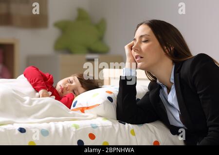 Müde Arbeiter Mutter Anzug schlafend neben schläfrig Tochter auf einem Bett zu Hause Stockfoto