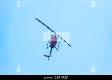 Ein kleiner zweisitziger kommerzieller Hubschrauber, der sich gegen einen klaren, blauen Himmel setzt, geht von unten nach oben. Ein kleiner Hubschrauber fliegt in einem klaren blauen Himmel. Stockfoto