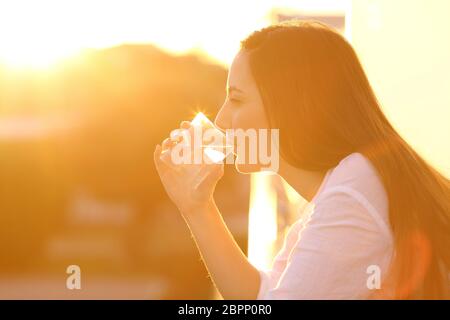 Seitenansicht Backlight Portrait einer Frau, die Wasser aus einem Glas in einem Haus Balkon bei Sonnenuntergang trinkt Stockfoto