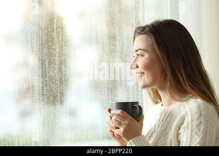 Gerne jugendlich Halten einer Tasse durch ein Fenster in einen regnerischen Tag suchen Stockfoto