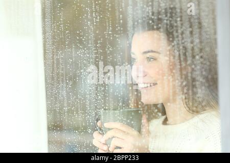 Smiley positive jugendlich Holding eine Kaffeetasse Suche im Freien durch ein Fenster in einen regnerischen Tag Stockfoto