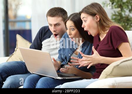 Drei Freunde, die Medieninhalte auf Linie in einem Computer sitzen auf einer Couch im Wohnzimmer zu Hause beobachten erstaunt Stockfoto