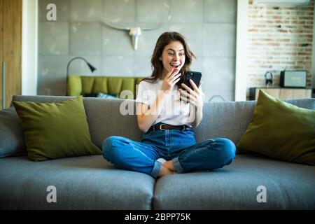 Porträt einer überraschten Frau, die ein Telefon hält und Sie im Wohnzimmer auf dem Sofa anschaut Stockfoto