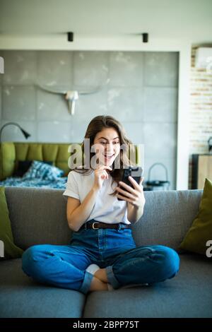 Porträt einer überraschten Frau, die ein Telefon hält und Sie im Wohnzimmer auf dem Sofa anschaut Stockfoto