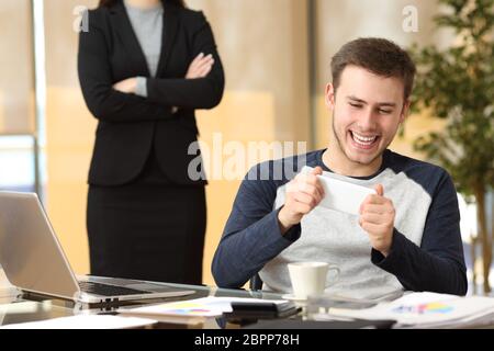 Faule Mitarbeiter spielen mit seinem Smartphone in einen Desktop-PC sitzen, während seine böse Chef im Büro zusieht Stockfoto