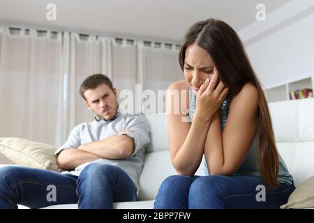 Frau, die häusliche Gewalt neben Ehemann wütend sitzen auf einer Couch im Wohnzimmer in einem Haus innen zu beklagen Stockfoto
