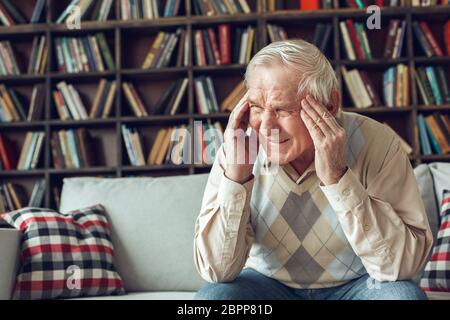 Der ältere Mann allein sitzt in der Bibliothek und hat schreckliche Kopfschmerzen Stockfoto