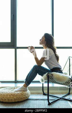 Junge Frau mit einer Tasse Getränk, die sich zu Hause im Schaukelstuhl entspannt Stockfoto