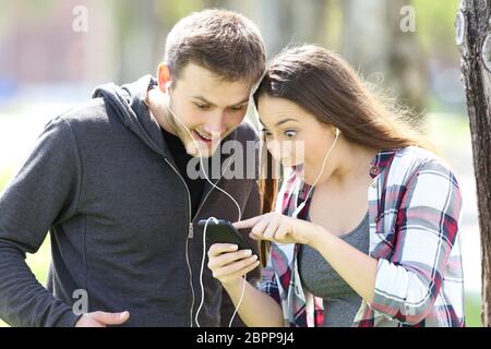 Erstaunt Paar ständigen hören die Musik auf Linie und Medieninhalte in einem Smart Phone im Freien in einem Park Stockfoto