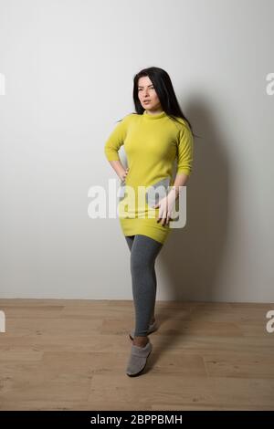 Ein Mädchen mit einem gelben langen Pullover und grauen Taschen in grauen Leggings steht auf einem grauen Wandhintergrund Stockfoto