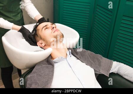 Friseur wäscht die Haare eines Kunden vor dem Schneiden. Ernst schöner Mann wäscht seinen Kopf in einem Schönheitssalon Stockfoto