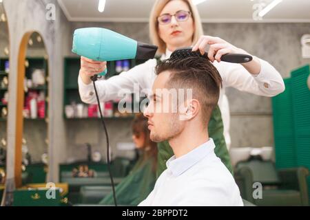 Friseur trocknet Haare von stilvollen Mann. Junge gutaussehende Kerl tun Haar Styling bei einem Friseur Stockfoto