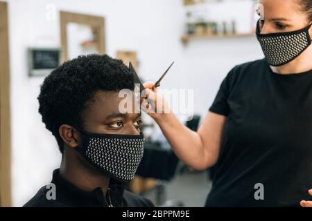 Gesicht eines schwarzen Mannes, der in einem Friseursalon einen Haarschnitt bekommt, mit einer schwarzen Maske auf seinem Gesicht vom Coronavirus. Der Friseur trägt auch eine Maske. Die Haare Stockfoto