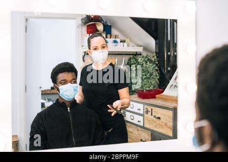 Schwarzer Kerl, der in einem Friseurladen seine Haare schneidet, mit einer schwarzen Maske auf seinem Gesicht vom Coronavirus. Der Friseur trägt auch eine Maske. Das Haar hat es Stockfoto