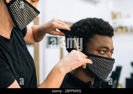 Gesicht eines schwarzen Mannes, der in einem Friseursalon einen Haarschnitt bekommt, mit einer schwarzen Maske auf seinem Gesicht vom Coronavirus. Der Friseur trägt auch eine Maske. Die Haare Stockfoto