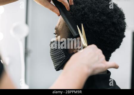 Gesicht eines schwarzen Mannes, der in einem Friseursalon einen Haarschnitt bekommt, mit einer schwarzen Maske auf seinem Gesicht vom Coronavirus. Das Haar ist wie bei Afro. Die Schere ist Gold Stockfoto