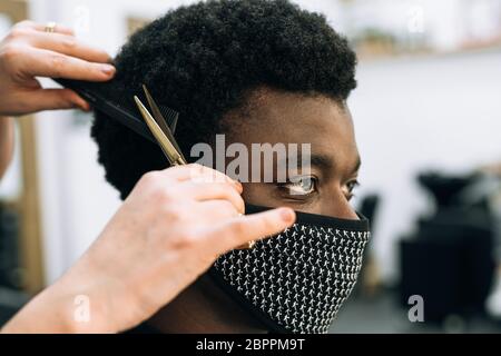 Gesicht eines schwarzen Mannes, der in einem Friseursalon einen Haarschnitt bekommt, mit einer schwarzen Maske auf seinem Gesicht vom Coronavirus. Das Haar ist wie bei Afro. Die Schere ist Gold Stockfoto