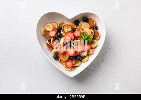 Trendy kleine Pfannkuchen mit Heidelbeere und Erdbeere in Teller mit Herz auf weißem Hintergrund geformt. Blick von oben. Stockfoto