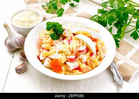 Salat von Crab Sticks, Käse, Knoblauch, Tomaten und Eier mit Mayonnaise in einer Platte, Handtuch, Petersilie und Gabel auf einem Holzbrett Hintergrund Stockfoto