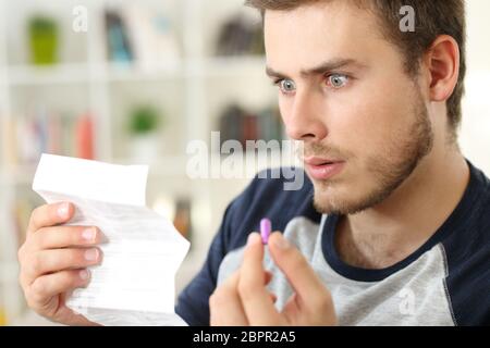 Schockiert Mann, der ein Flugblatt liest, bevor er eine rosa Pille nimmt, die auf einem Sofa im Wohnzimmer in einem Hausinneren sitzt Stockfoto