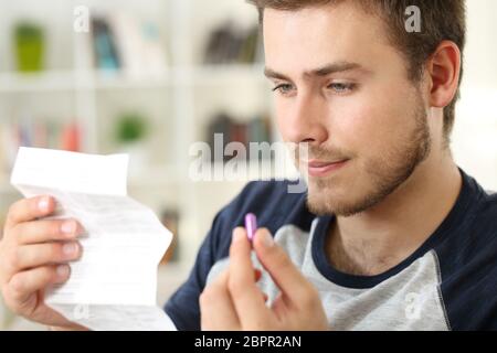 Mann, der ein Flugblatt liest, bevor er eine rosa Pille nimmt, die auf einem Sofa im Wohnzimmer in einem Hausinneren sitzt Stockfoto