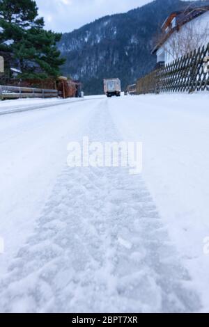 Weitwinkel- und bodengleiche Ansicht entlang einer schneebedeckten Dorfstraße mit einer Reifenspur links vom LKW, der zum Fahrzeug führt. Stockfoto