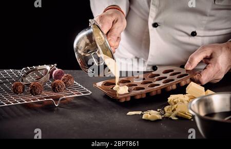 Koch oder Chocolatier gießen geschmolzene weiße Schokolade in Silikon Backformen aus einem Topf in einer Nahaufnahme auf seinen Händen Stockfoto