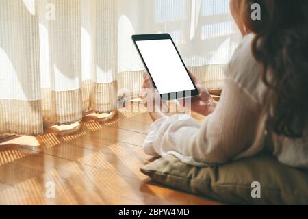 Mockup-Bild einer Frau, die schwarzen Tablet-pc mit leeren weißen Desktop-Bildschirm hält, während sie sich auf dem Boden mit Gefühl entspannt Stockfoto