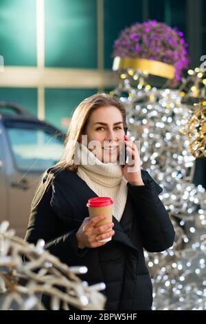 Porträt beschäftigt moderne hübsche Frau im Gespräch auf Smartphone. Attraktive Geschäftsfrau hält Kaffee zum Mitnehmen, mit dem Telefon. Geschäftsfrau im Hintergrund modernes Business-Center. Bokeh weihnachtsbeleuchtung Stockfoto