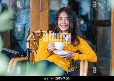 Asiatische junge Frau, die eine Tasse Kaffee hält und im modernen Café oder Coworking Space neben dem Fensterspiegel sitzt und Kopfhörer zum Hören trägt Stockfoto