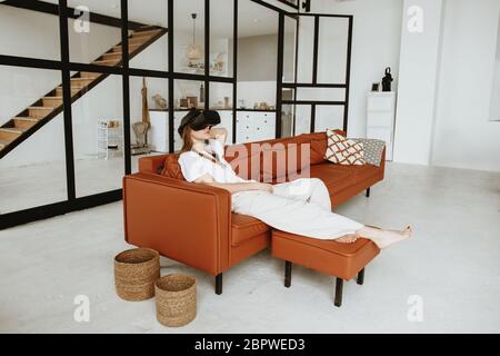 Schöne junge Frau sitzt auf dem Sofa zu Hause mit Virtual Reality-Headset. Stockfoto