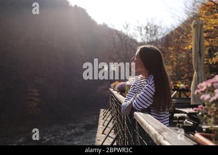 Junge schöne Frau genießt den Blick auf den Bergfluss und zeigt die Zunge auf der Terrasse eines Cafés an einem sonnigen Herbsttag. Selektive Fokussierung Stockfoto