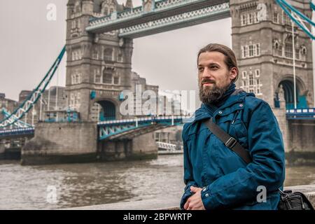 Ein junger Tourist mit langem Haar und dickem Bart steht im Winter bei bewölktem Wetter auf dem Hintergrund der London Bridge Stockfoto