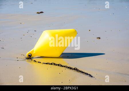 Eine große gelbe Boje und ihre Ankerkette, die als Abschusskanallinder verwendet wird, liegen bei Ebbe am Strand in der Sonne auf dem nassen Sand. Stockfoto