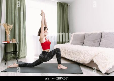 Eine Frau stürzt sich während der Quarantäne und Selbstisolation zu Hause die Beine. Fitness-Mädchen in athletischen Kleidung tun vorwärts Ausfallschritt Training Beinmuskeln in fla Stockfoto