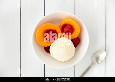 Draufsicht auf einen Pfirsichmelba-Sonnenteller in einer weißen Porzellanschüssel gefüllt mit Vanilleeis, Pfirsichen, Himbeeren und Himbeersauce auf weißer Holzplatte Stockfoto