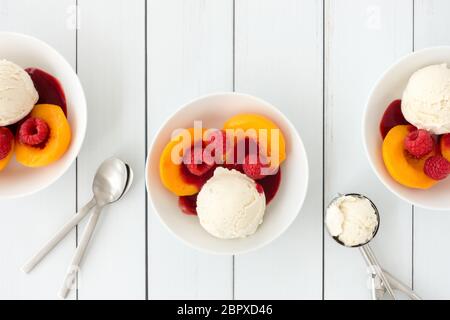 Schalen gefüllt mit Vanilleeis, Pfirsiche, Himbeeren und Himbeersauce auf weißem Holztafel Hintergrund mit Kopierraum. Draufsicht. Stockfoto