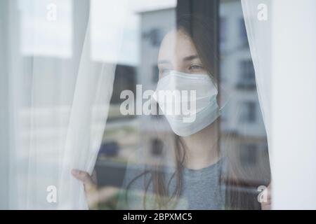 Junge Frau im Gesicht Maske Blick aus dem Fenster. Wohnen zu Hause in der Selbstquarantäne. Stockfoto