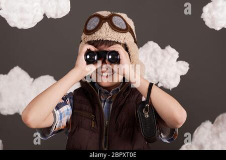 Ein Junge spielt mit Ferngläsern und träumt davon, Pilot zu werden, gekleidet in eine Retro-Jacke und Helm mit Brille, Wolken aus Watte, grauer Rücken Stockfoto