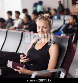 Casual braungebrannte blonde Frau mit Handy, Reisepass und Bordkarte während der Wartezeit in ein Flugzeug steigen an den Abfluggates an den asiatischen zu Stockfoto
