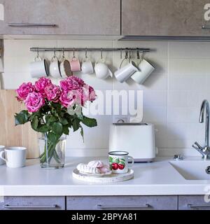 Küche in beigefarbenen Tönen. Toaster. Becher und Milchkännchen hängen an Haken. Waschbecken mit Wasserhähnen. Ein Tablett mit Becher und Marshmallows. Bouquet von roten Rosen in einem Glas Stockfoto