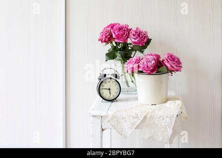 Ein Strauß rosa Rosen in einem Zinn weißen Eimer auf einem alten Hocker mit Wecker und einer Serviette mit Stickerei neben dem Bett auf einem beigen Hintergrund Stockfoto