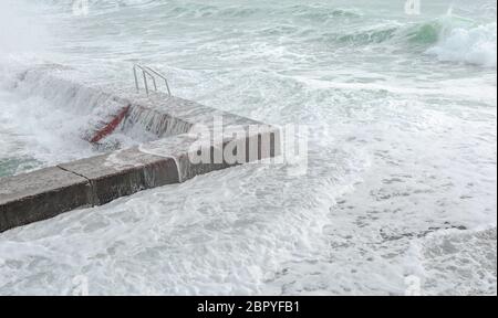 Wellen schlagen auf Wellenbrechern mit roten Treppen und verwandeln sich in Milchschaum Stockfoto