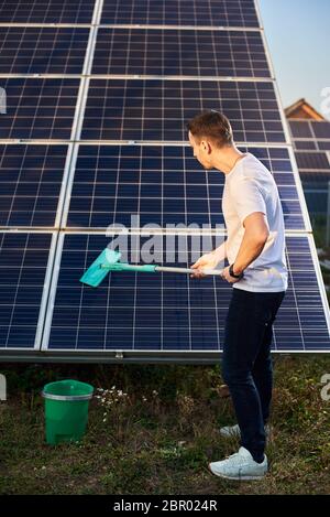 Man reinigt Solarpanel in Solarkraftwerk. Der junge Mann ist stilvoll gekleidet mit einer Uhr an seinen Händen auf einem Grundstück in der Nähe des Hauses Stockfoto