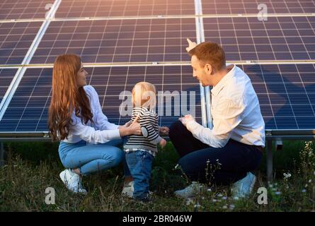 Vater zeigt seiner Familie die Sonnenkollektoren auf dem Grundstück in der Nähe des Hauses an einem warmen Tag. Ein junges Mädchen mit einem Kind und ein Mann in den Sonnenstrahlen schauen sich die Sonnenkollektoren an. Stockfoto