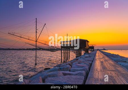 Traditionelles Fischerstationshaus mit Netz im Wasser der Adria am Pier Diga Sottomarina, Skyline mit Strand, erstaunlicher gelb-roter Sonnenuntergang in der Dämmerung, Dämmerung, Abendansicht, Norditalien Stockfoto