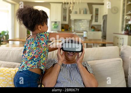 Ältere Mischrassenfrau mit ihrer jungen Enkelin, die ihr dabei hilft, ein VR-Headset anzuziehen Stockfoto