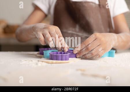 Ein kleines Mädchen trägt eine braune Schürze mit einer lila Form, um den Teig für die Herstellung von Cookies in der Küche zu schneiden. Stockfoto