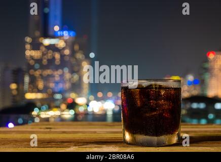 Nahaufnahme eines Glases kohlensäurehaltigen Erfrischungsgetränks oder einer Cola mit Eis und Nachtansicht der Bokeh-Lichter der Stadt Stockfoto