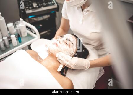 Der Kosmetologe wäscht die Maske vom Gesicht des Patienten ab. Entfernen einer Feuchtigkeitsmaske aus dem Gesicht Stockfoto